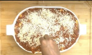 Hand sprinkling cheese on oven-baked pasta dish in a white casserole dish on a wooden board.